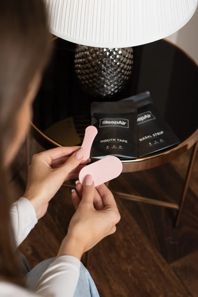 Kézben tartott SleepAir tapaszok, háttérben Mouth Tape és Nasal Strip csomagolással